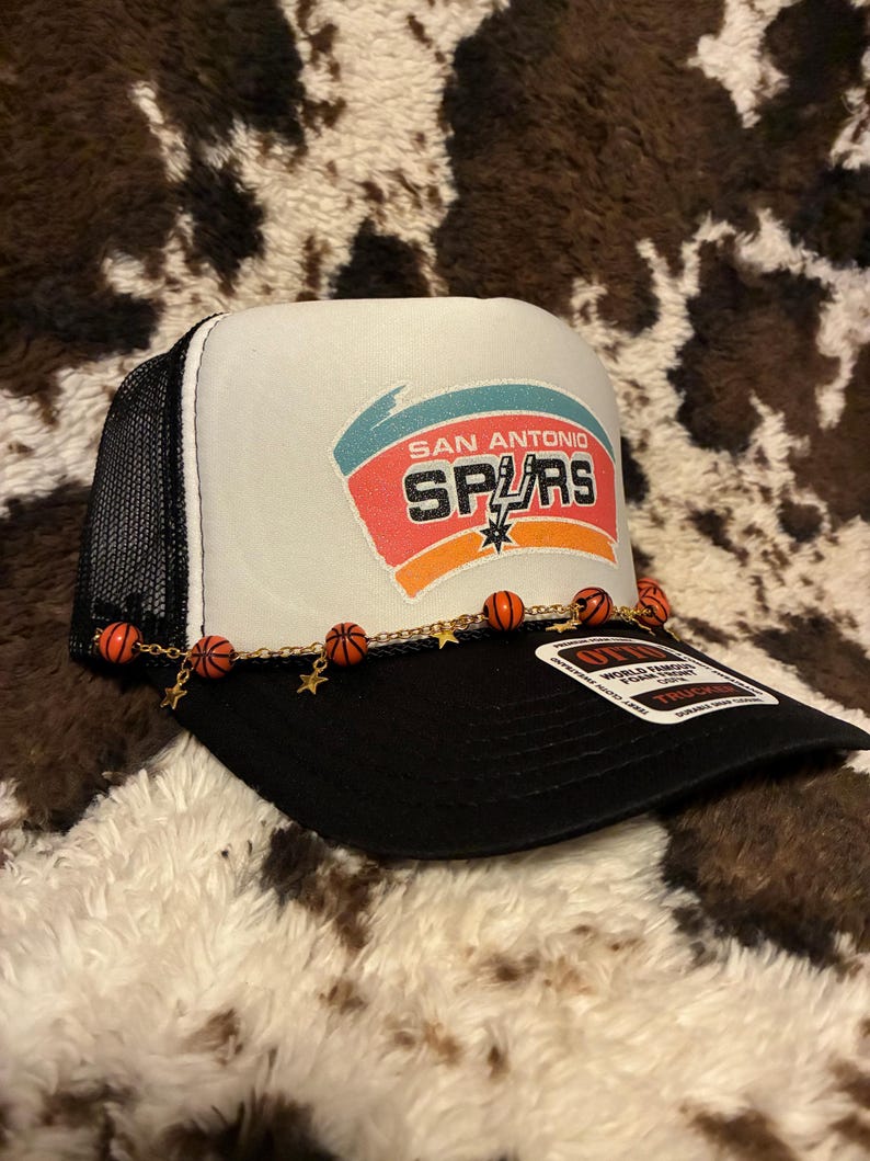 May include: A black and white trucker hat with the San Antonio Spurs logo on the front. The hat features a mesh back, a curved brim, and a decorative chain with basketball and star charms.
