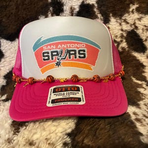 May include: A pink and white trucker hat with the San Antonio Spurs logo. The hat features a white front panel with the team's logo in teal, orange, and black. A string of small basketballs decorates the brim. The hat has a pink mesh back.