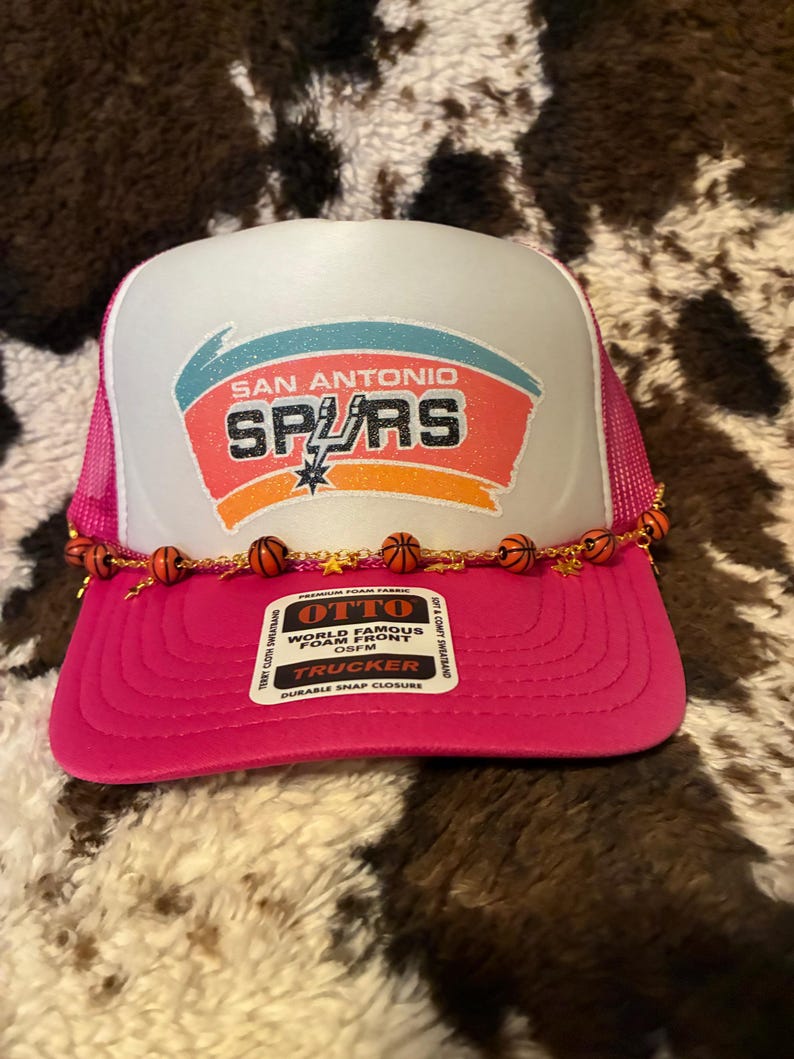 May include: A pink and white trucker hat with the San Antonio Spurs logo. The hat features a white front panel with the team's logo in teal, orange, and black. A string of small basketballs decorates the brim. The hat has a pink mesh back.