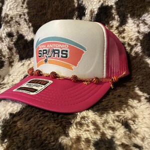 May include: A pink and white trucker hat featuring the San Antonio Spurs logo. The hat has a mesh back and a curved brim. A gold chain with basketball charms and star charms is attached to the brim.