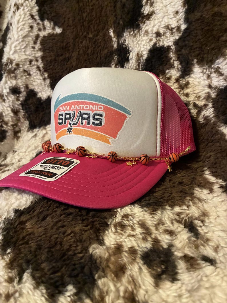 May include: A pink and white trucker hat featuring the San Antonio Spurs logo. The hat has a mesh back and a curved brim. A gold chain with basketball charms and star charms is attached to the brim.