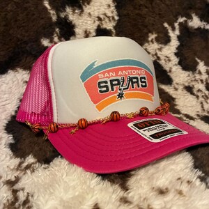May include: A pink and white trucker hat featuring the San Antonio Spurs logo. The hat has a pink mesh back and a curved brim. A decorative chain with basketball charms adorns the brim. The hat is on a cowhide-patterned surface.