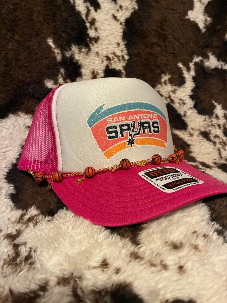 May include: A pink and white trucker hat featuring the San Antonio Spurs logo. The hat has a pink mesh back and a curved brim. A decorative chain with basketball charms adorns the brim. The hat is on a cowhide-patterned surface.