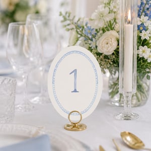 May include: An elegant table setting with a white tablecloth, featuring a table number card with the number 1 in blue, surrounded by a blue dotted border. A gold-colored metal holder supports the card. The setting includes glassware, flowers, and a lit candle.