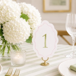 May include: A white table number card with a scalloped edge, a light pink border, and the number 1 printed in a soft green watercolor style. The card is held upright by a small, circular gold metal stand on a striped green and white tablecloth.