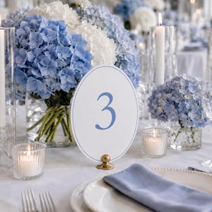 May include: A table setting with blue and white hydrangeas, candles, and a table number. The centerpiece features a large bouquet of blue and white hydrangeas in a glass vase. A white oval table number with the number 3 in blue is displayed.