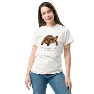 Puede incluir: Una camiseta blanca de manga corta con un gráfico realista de una tortuga marrón centrado en el pecho. Debajo de la tortuga, el texto dice "slow motion over no motion" en una fuente serif negra simple. La camiseta tiene cuello redondo y corte estándar.