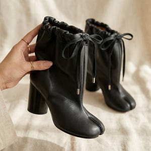 May include: A pair of black leather ankle boots featuring a split-toe design, a gathered drawstring collar with a tied bow, and a sturdy block heel. The boots have a smooth finish and a vertical seam running down the front center of the shaft.