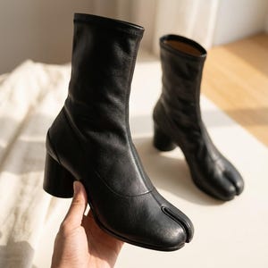May include: A pair of black ankle boots featuring a split-toe design, a mid-height block heel, and a smooth, matte finish. The boots have a fitted, sock-like shaft that extends above the ankle, with a vertical seam running down the front center.