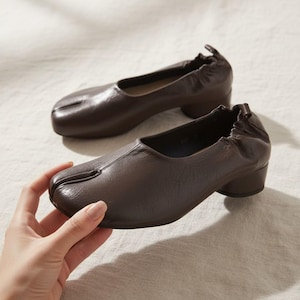 Op de afbeelding: Een paar donkerbruine leren schoenen met een split-teenontwerp. De schoenen hebben een lage, afgeronde hak en een geplooid detail aan de achterkant. De schoenen zijn ongeveer 5 cm hoog.