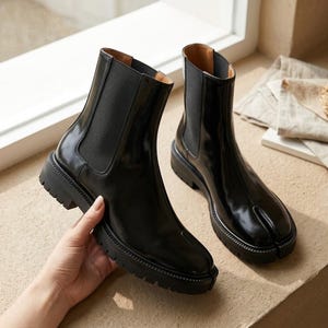 May include: A pair of black ankle boots with a glossy finish, featuring a split-toe design, elastic side panels, and a thick, lugged rubber sole. The boots have a structured silhouette with a vertical seam running down the center of the toe box.