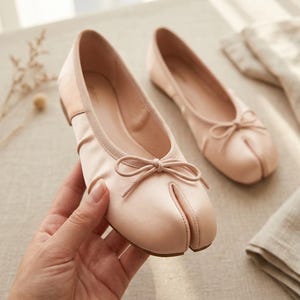 May include: A pair of pale pink ballet flats featuring a split-toe design and a small decorative bow on the front. The shoes have a soft, smooth finish and a low-profile silhouette, with one shoe held up to show the detail of the toe construction.