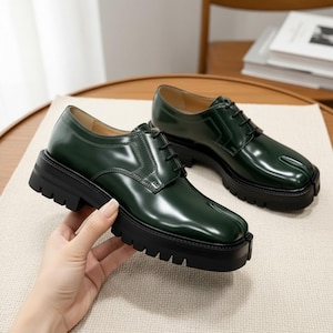 May include: A pair of dark green leather shoes with black soles. The shoes have a lace-up design and a unique split-toe detail. The thick, black soles add a chunky, modern look. The shoes are displayed on a neutral surface.