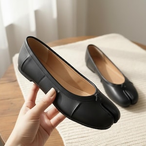 Pode incluir: Sapatilhas de couro preto com design de bico dividido. Os sapatos têm um pequeno laço na frente e um bico arredondado. O interior é de cor bege claro. Um sapato é segurado por uma mão, o outro repousa sobre uma superfície creme.