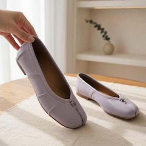 May include: A pair of lavender ballet flats with a square toe and small decorative bow. The shoes have a smooth, leather-like appearance and a low heel. One shoe is being held by a hand, while the other rests on a light-colored surface.
