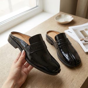 May include: A pair of black patent leather slip-on loafers featuring a split-toe design, a low block heel, and a classic penny loafer strap across the vamp. The shoes have a glossy finish and an open-back construction for easy wear.