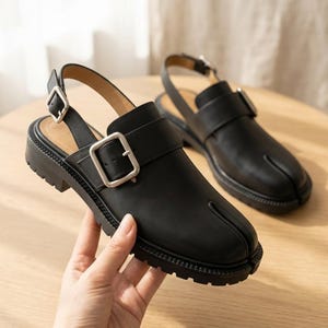 May include: A pair of black leather slingback sandals featuring a split-toe design, a thick lug sole, and a prominent silver-toned metal buckle on the side strap. The shoes have a smooth finish and a sturdy construction with an adjustable heel strap.