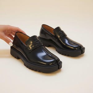 Op de afbeelding: Paar zwarte leren loafers met een split-teen ontwerp. De schoenen hebben een dikke, profielzool en een slip-on stijl met een decoratieve band. De binnenkant is lichtbruin.