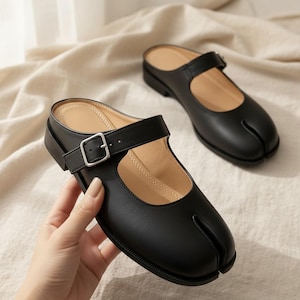 May include: A pair of black split-toe mules featuring a Mary Jane style strap with a silver-toned metal buckle. The shoes have a smooth finish, a low heel, and a beige interior lining. The design includes a distinct cleft at the front toe area.