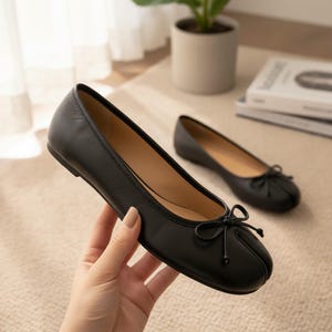 Könnte beinhalten: Schwarze Ballerinas aus Leder mit einer kleinen Schleife auf der Spitze. Die Schuhe sind flach und haben eine runde Spitze. Ein Schuh wird in einer Hand gehalten, ein anderer befindet sich im Hintergrund. Die Schuhe sind ein klassischer Stil.
