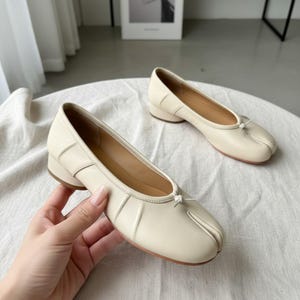 Peut inclure: Ballerines en cuir blanc cassé avec un design à bout fendu. Les chaussures ont un petit talon et un détail de nœud sur le dessus. Les ballerines sont sur une surface blanche.