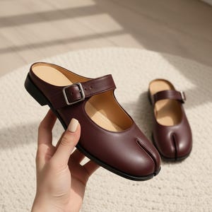 May include: A pair of dark burgundy slip-on shoes featuring a split-toe design, a low heel, and a single adjustable strap across the top with a silver-toned metal buckle. The interior of the shoe is lined in a light beige material.