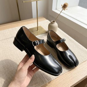 May include: A pair of black leather Mary Jane shoes featuring a distinctive split-toe design, a low block heel, and a thin adjustable strap across the instep secured with a small silver-toned metal buckle. The shoes have a smooth finish.