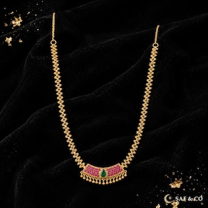 May include: A gold-toned necklace featuring a textured chain and a central curved pendant. The pendant is inlaid with small pink stones and a single central teardrop-shaped green stone, finished with a row of small dangling gold beads at the base.