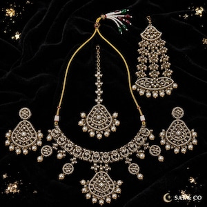 May include: A jewelry set featuring a necklace, two earrings, a maang tikka, and a jhoomar, all crafted with intricate silver-toned metalwork, clear crystals, and small white pearls. The necklace has a central pendant and adjustable cord.