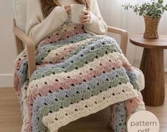May include: A chunky crochet blanket featuring alternating horizontal stripes in cream, dusty pink, and sage green. The textured yarn creates a repeating shell-like pattern throughout the fabric. A circular beige graphic displays the text pattern pdf.