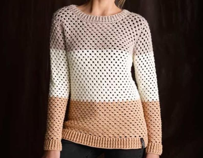 May include: A long-sleeved crochet sweater featuring a color-blocked design with horizontal stripes in shades of light beige, white, and tan. The garment has a round neckline and ribbed cuffs and hem, constructed with a repeating openwork stitch.