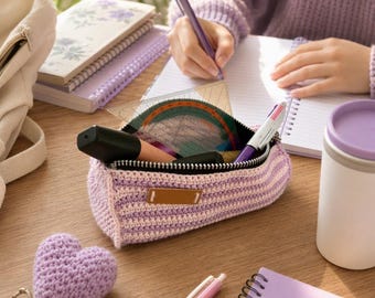 May include: A crochet pencil case with horizontal light purple and white stripes, featuring a silver zipper and a rectangular brown leather-like label. The case contains a clear plastic protractor, a black marker, and a multi-colored pen.