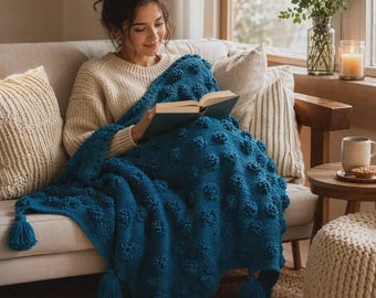 May include: A deep teal crochet throw blanket featuring a textured bobble stitch pattern and large tassels at the corners. The blanket is draped over a beige sofa, covering a person who is holding an open book with a dark blue cover.