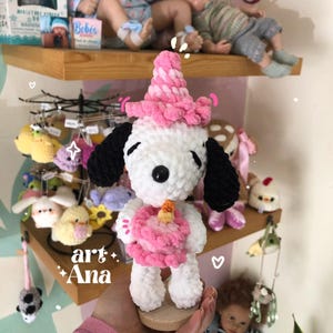以下が含まれることがあります： 黒い耳を持つビーグル犬に似た白いかぎ針編みのぬいぐるみ。ピンクのパーティーハットをかぶり、オレンジ色のキャンドルが乗った小さなピンクのバースデーケーキを持っています。ぬいぐるみは柔らかい質感の毛糸でできており、小さな木製の台座の上に立っています。