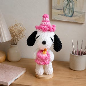 以下が含まれることがあります： 黒い耳を持つ白いかぎ針編みの犬のフィギュア。ピンクのパーティーハットをかぶり、黄色いキャンドルがのった小さなピンクのバースデーケーキを持っています。このぬいぐるみは円形の木製台座の上に立っており、黒い鼻と閉じた目があります。