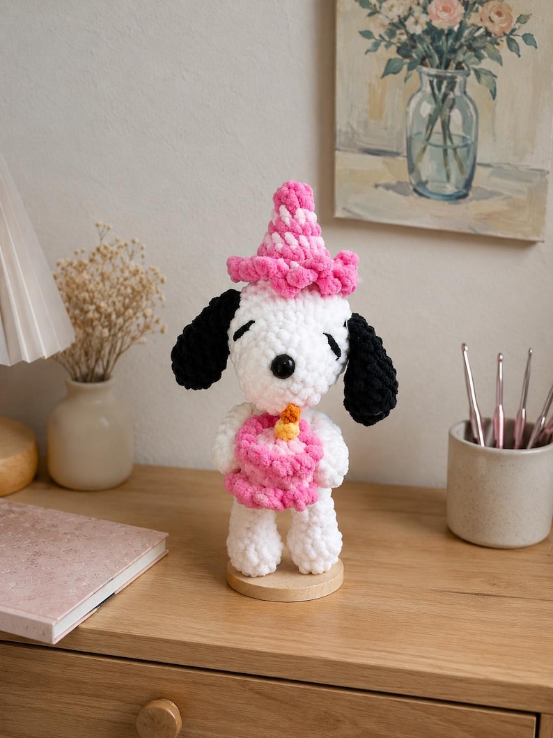 以下が含まれることがあります： 黒い耳を持つ白いかぎ針編みの犬のフィギュア。ピンクのパーティーハットをかぶり、黄色いキャンドルがのった小さなピンクのバースデーケーキを持っています。このぬいぐるみは円形の木製台座の上に立っており、黒い鼻と閉じた目があります。