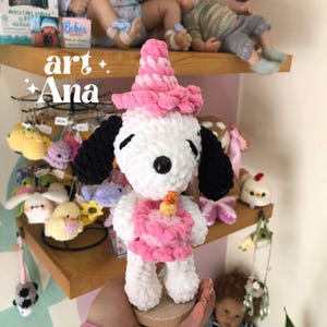 以下が含まれることがあります： ピンクのパーティーハットをかぶり、ろうそくのついた小さなピンクのケーキを持っている、白と黒の編みぐるみの犬のフィギュア。黒い耳、黒い鼻、閉じられた目を持つ。左上に「art Ana」という文字が見える。