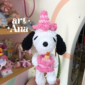 以下が含まれることがあります： 黒い耳を持つ白いかぎ針編みのぬいぐるみ犬。ピンクのパーティーハットをかぶり、黄色いキャンドルが乗った小さなピンクのかぎ針編みのバースデーケーキを持っています。柔らかい毛糸で作られ、小さな木製の台座の上に立っています。