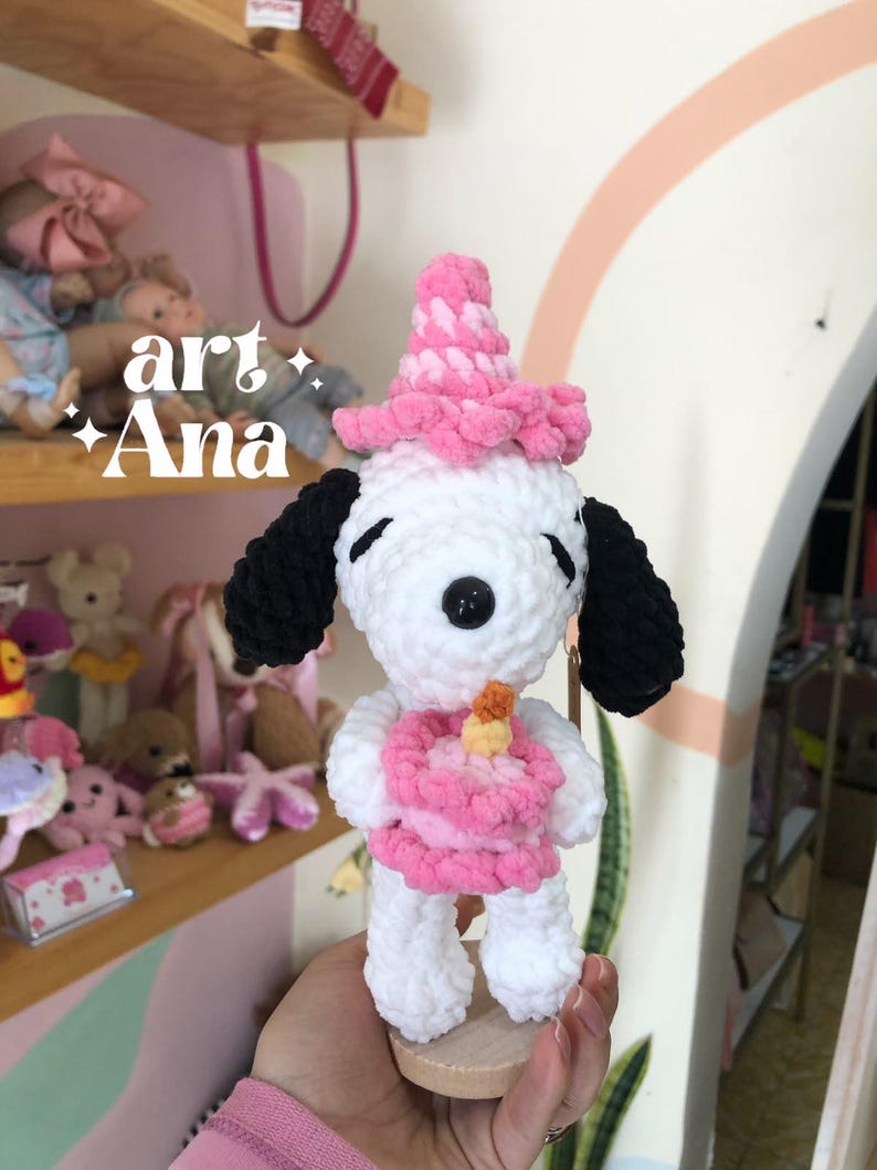 以下が含まれることがあります： 黒い耳を持つ白いかぎ針編みのぬいぐるみ犬。ピンクのパーティーハットをかぶり、黄色いキャンドルが乗った小さなピンクのかぎ針編みのバースデーケーキを持っています。柔らかい毛糸で作られ、小さな木製の台座の上に立っています。