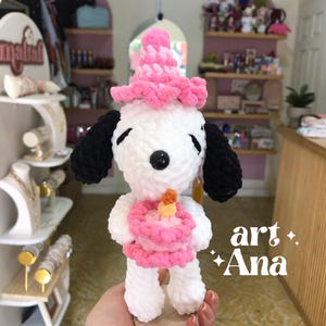 以下が含まれることがあります： 黒い耳を持つ白いかぎ針編みのぬいぐるみ犬。ピンクのパーティーハットをかぶり、黄色いキャンドルが乗った小さなピンクのケーキを持っています。ぬいぐるみには黒い鼻と閉じた目があります。右下に「art Ana」という文字が見えます。