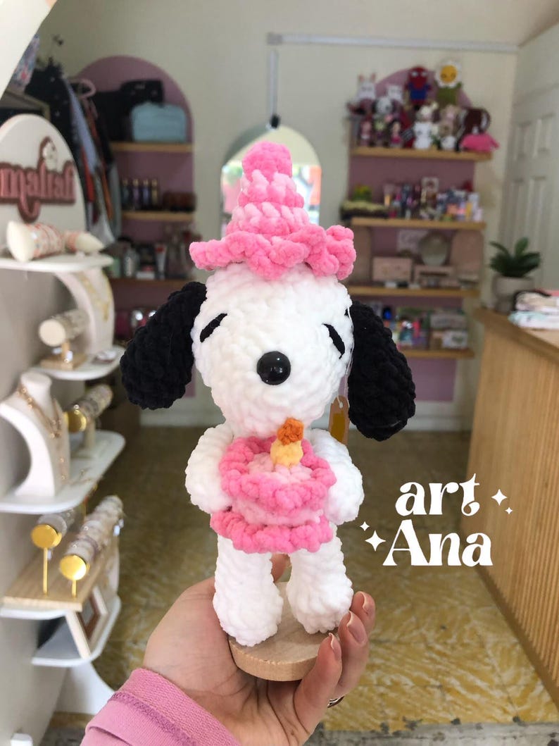 以下が含まれることがあります： 黒い耳を持つ白いかぎ針編みのぬいぐるみ犬。ピンクのパーティーハットをかぶり、黄色いキャンドルが乗った小さなピンクのケーキを持っています。ぬいぐるみには黒い鼻と閉じた目があります。右下に「art Ana」という文字が見えます。