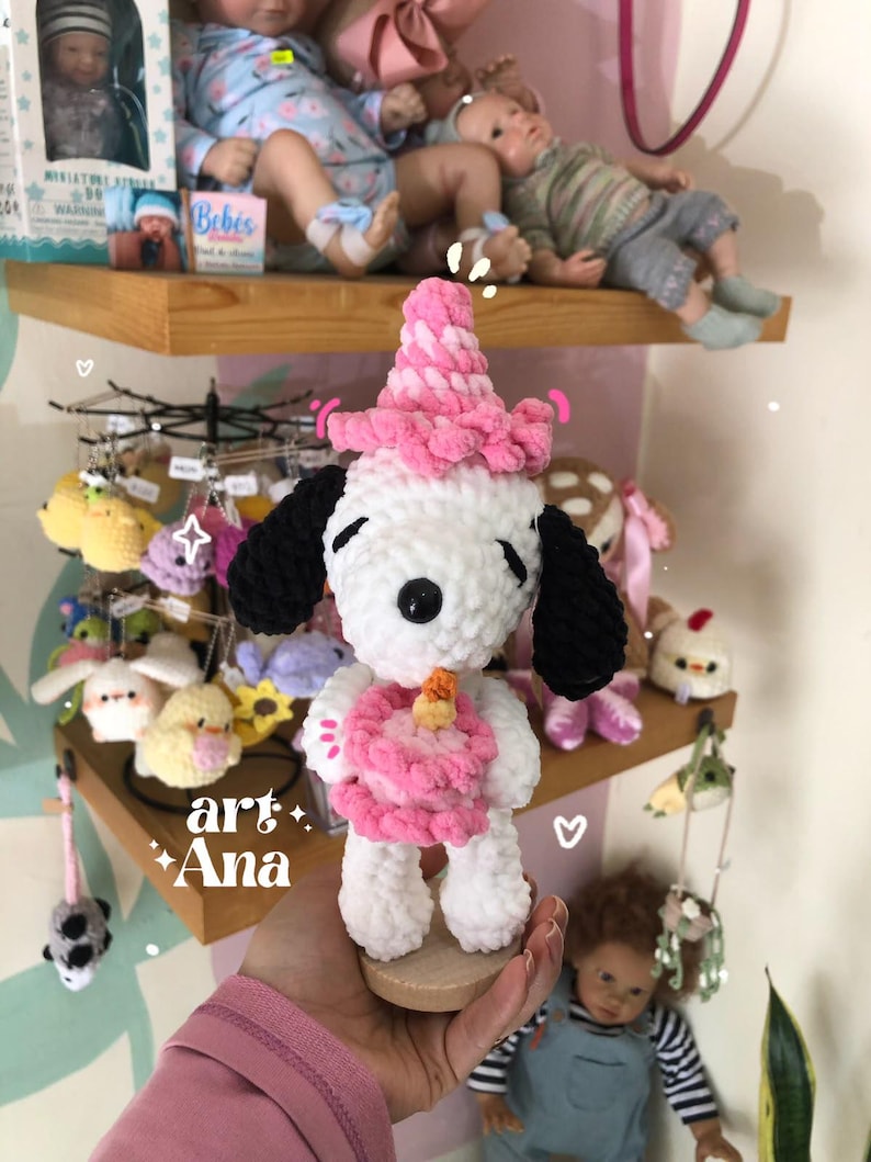 以下が含まれることがあります： 黒い耳を持つビーグル犬に似た白いかぎ針編みのぬいぐるみ。ピンクのパーティーハットをかぶり、オレンジ色のキャンドルが乗った小さなピンクのバースデーケーキを持っています。ぬいぐるみは柔らかい質感の毛糸でできており、小さな木製の台座の上に立っています。