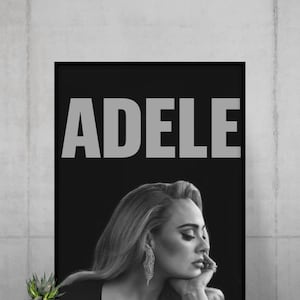 May include: A black and white framed poster featuring the name ADELE in large bold capital letters above a portrait of the singer. The portrait shows a side profile of the individual with long hair, a dangling earring, and a hand near the chin.