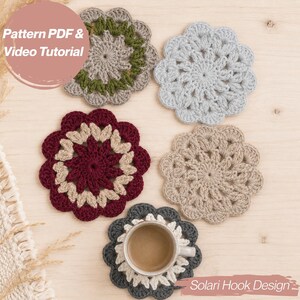 May include: Five round crochet coasters with scalloped edges in various colors including burgundy, beige, light blue, and dark gray. One dark gray coaster supports a ceramic mug filled with coffee. Text reads Pattern PDF & Video Tutorial.