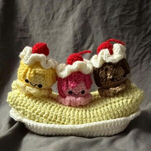 May include: A crocheted banana split with three scoops of ice cream. The ice cream scoops are yellow, pink, and brown, each topped with a red cherry. The banana boat is yellow and white.
