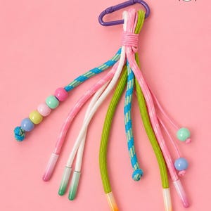 May include: A vibrant lanyard with a purple carabiner clip. It features cords in pink, green, white, and blue, adorned with beads and gradient-coloured tips. The background is a solid pink. A cartoon illustration of a girl is in the top right corner.