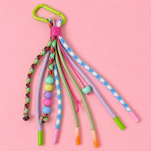 Peut inclure: Un porte-clés coloré avec un mousqueton vert. Il présente plusieurs cordons de différentes couleurs et motifs, dont un cordon à motifs arc-en-ciel, un cordon à carreaux bleu clair et blanc, et des cordons roses et violets unis. Les cordons sont ornés de perles et finis avec des embouts colorés.