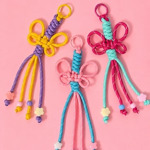 May include: Three colourful handmade keychains with a floral design. Each keychain features a braided knot with a flower-shaped design and dangling cords. The keychains are in shades of yellow, purple, pink, blue, and green, with decorative beads and flower charms.