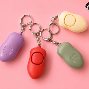 May include: Four personal safety alarms in purple, red, yellow, and green, each attached to a silver keychain. The purple alarm has a button, while the red one has a circular activation area. The green alarm has "SOS" printed on it. All are on a pink background.
