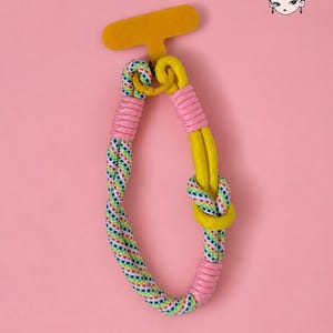 Könnte beinhalten: Ein farbenfroher Handy-Gurt mit gelbem Tab und geflochtenem Seildesign. Der Gurt hat eine Kombination aus rosa, gelbem und mehrfarbigem Seil und schafft ein lebendiges Accessoire. Der Hintergrund ist einfarbig rosa.